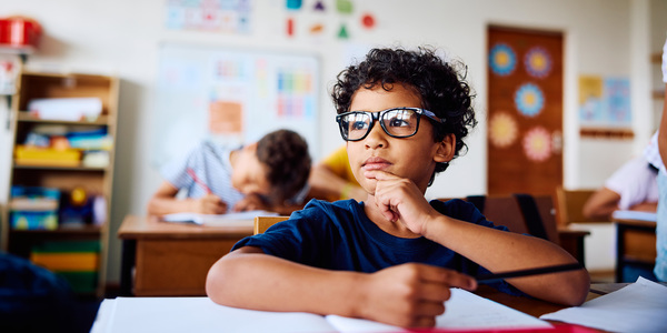 Jongen met bril zit aan een lessenaar in een klaslokaal en kijkt mijmerend naar buiten.